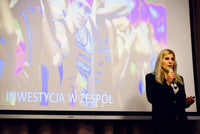 Kobieta, blondynka ubrana w garnitur stoi przy ekranie z mikrofonem w ręku i przemawia do ludzi. W tle towarzyszy jej wyświetlona prezentacja.